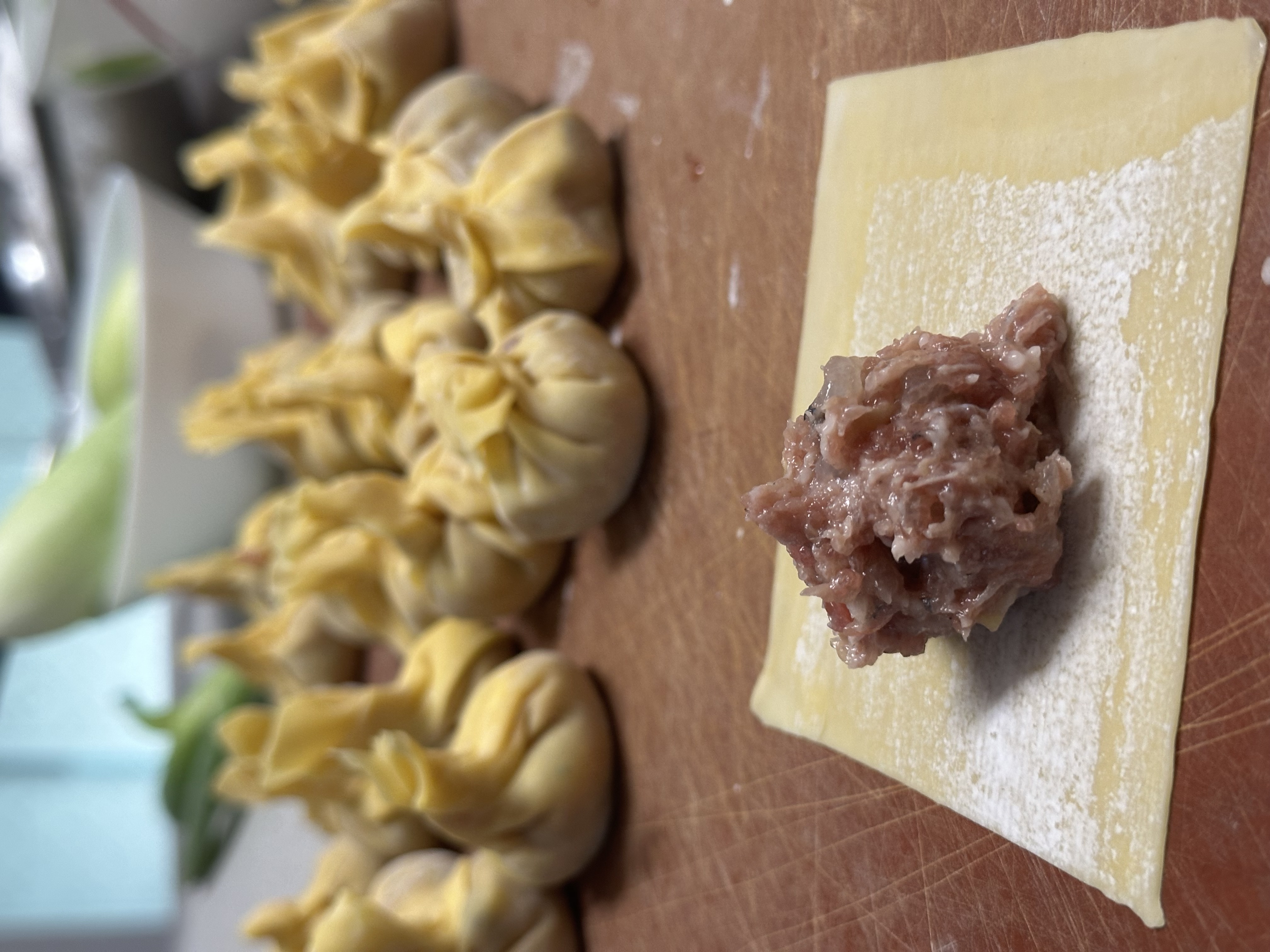 Photo of a wad of dumpling filling sitting on top of a flat square of wonton wrapper. Several finished dumplings just out of focus in the background 